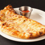A delicious handmade brick oven Calzone pizza sandwich with marinara sauce composed on a simple black background.